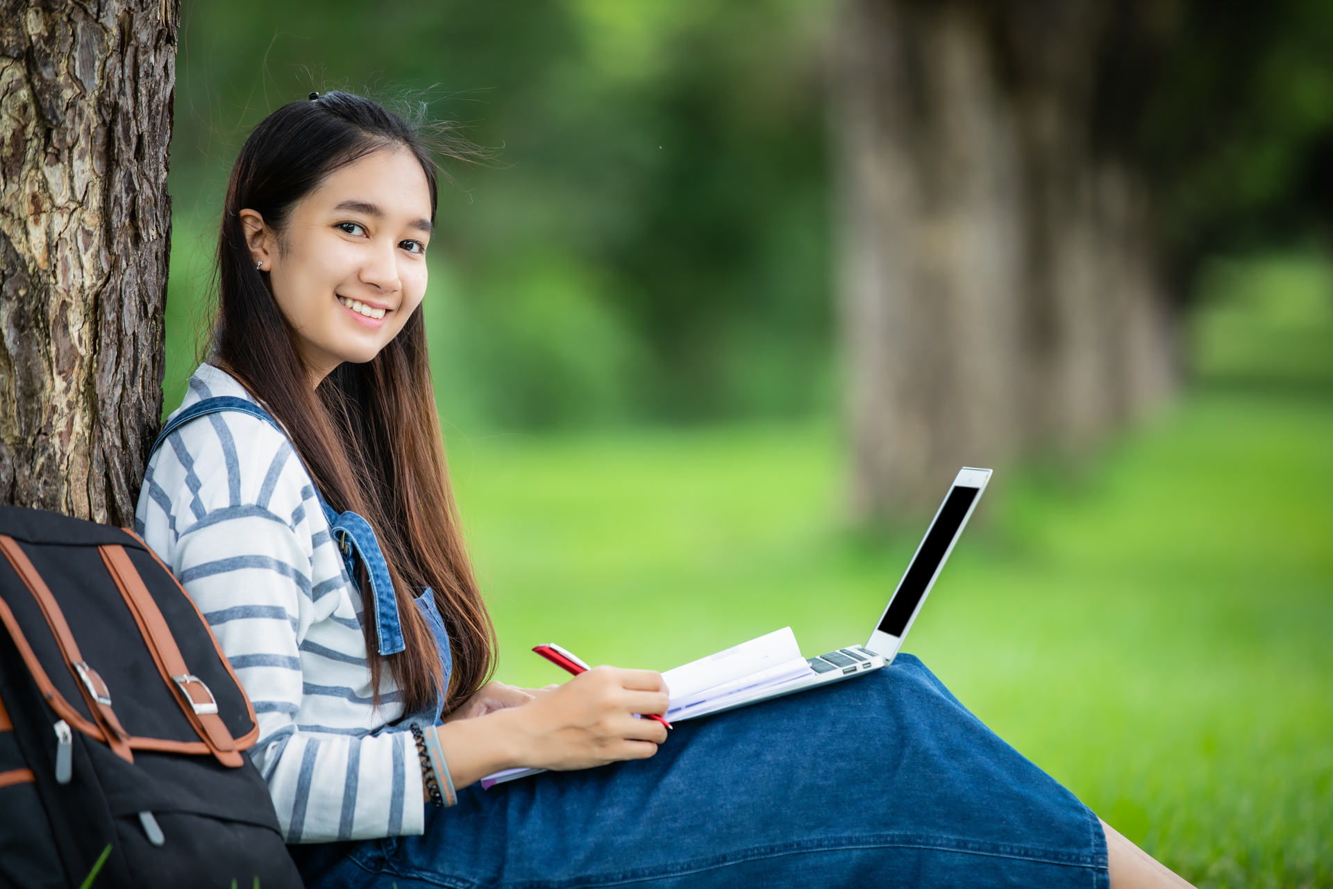 Studentka UWM siedząca pod drzewem z laptopem na kolanach